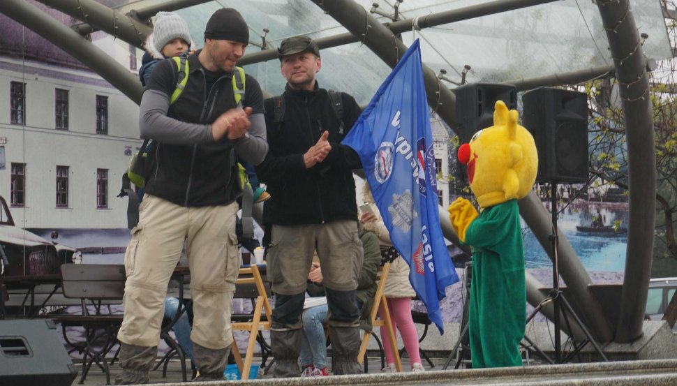 Obrazek przedstawia stojących na scenie na rynku w Ustronie dwóch nieumundurowanych policjantów, trzymających jasia w foteliku na plecach, którzy przeszli ponad 500 km po górach, na obrazku widoczne także inne postacie w tle