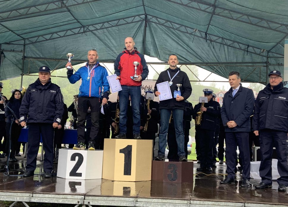 Obrazek przedstawia stojących z pucharami i dyplomami na podium nieumundurowanych policjantów, obok podnium po obu stronach stroją umundurowani policjanci  za nimi inne osoby