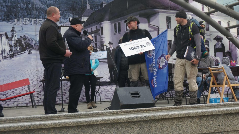 Obrazek przedstawia czterech mężczyzn na scenie, jeden z nich na plecach trzyma w foteliku chłopca, obok niego inny tzryma flagę "Wyprawa dla Jasia oraz potwierdzenie zapłaty w postaci bonu, jedne z mężczyzn po lewej stronie przemawia przez mikrofon lewej stronie ny mężczyzny 