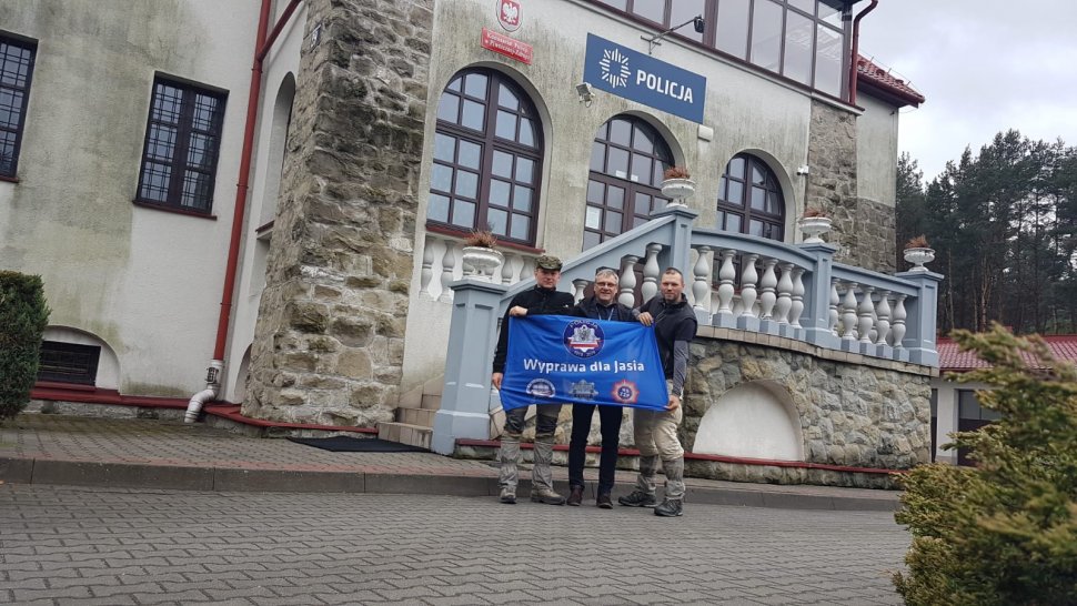 Obrazek przedsatwia trzech mężczzyn trzymających flagę "Wyprawa dla Jasia" na tle komendy Policji 