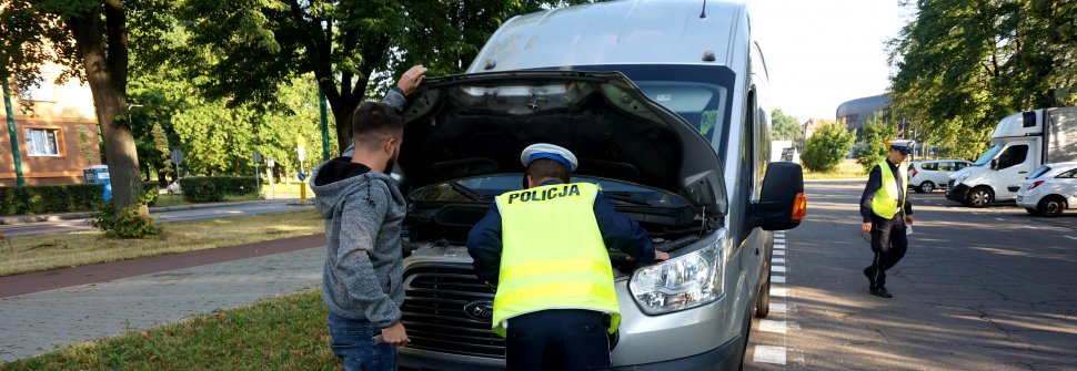 Policjant zagląda pod maskę, kierowca trzyma klapę, drugi policjant widoczny w tle. 