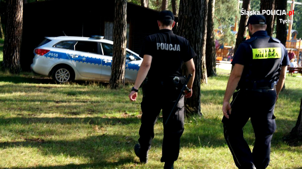 Na zdjęciu widoczny tyłem policjant oraz strażnik miejski, obaj idą w stronę radiowozu. 