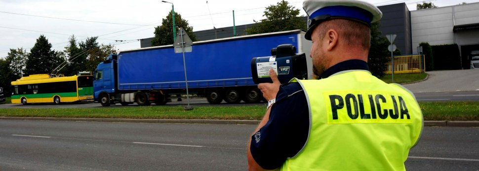 Na zdjęciu widoczny policjant wydziału ruchu drogowego w kamizelce odblaskowej stoi tyłem na drodze, w tle widoczny tir i autobus.