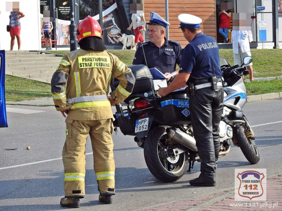 Dwóch policjantów stoi przy policyjnym motorze jeden stoi przodem drugi tyłem obok tyłem stoi funkcjonariusz straży pożarnej.