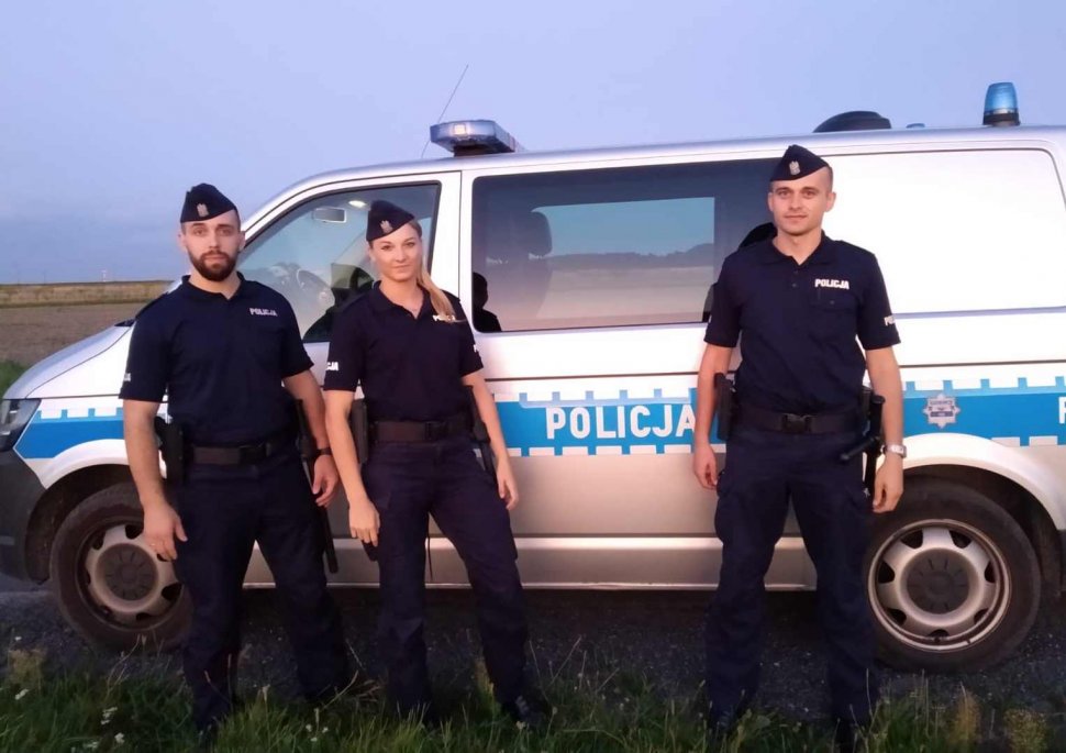 Trzech umundurowanych policjantów ze Szkoły Policji w Katowicach  w  środku policjantka) stoi przed oznakowanym radiowozie. 