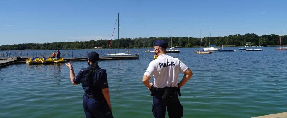 Policjanci stoją nad brzegiem zbiornika &quot;Paprocany&quot;, w tle widoczne są żaglówki i rowerki wodne.