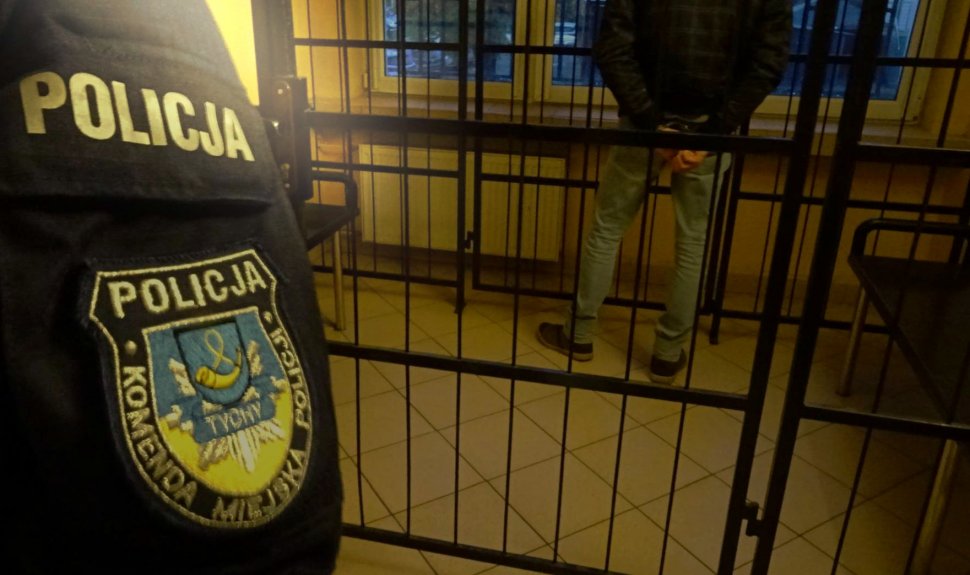 Za kratkami aresztu stoi zatrzymany mężczyzna, w tle widoczny mundur policjanta z naszywką Komendy Miejskiej Policji w Tychach. 