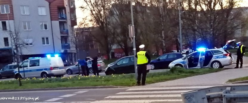 Na zdjęciu widoczny policjant, który stoi tyłem na chodniku, obok inne pojazdy oraz radiowóz z włączonymi sygnałami świetlnymi.
