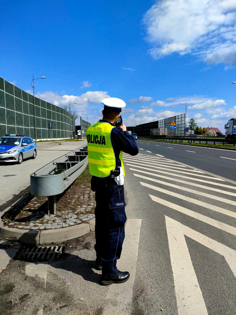 Umundurowany policjant mierzy w kierunku samochodów ręcznym miernikiem prędkości i mierzy prędkość pojazdów. 