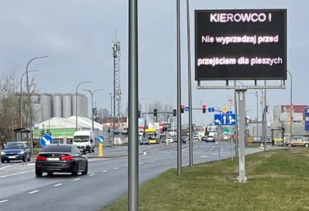 Na zdjęciu widoczna elektroniczna tablica przy drodze z wyświetlanym komunikatem o treści "Kierowco! Nie wyprzedzaj przed przejściem dla pieszych!"