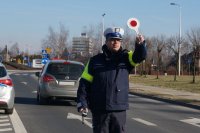 Obrazek przedstawia stojącego na jezdni umundurowanego policjanta z tarczą do zatrzymywania kierowców, za nim w tle odjeżdżający samochód