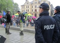 Obrazek przedstawia strojących na scenie nieumundurowanych policjantów, jedne z nich w foteliku na plecach trzyma chłopca, po prawej stronie (tyłem) stoi dwóch umundurowanych policjantów-Komendant z Tychów i z Cieszyna