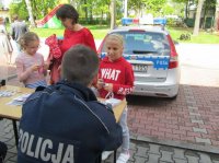 Obrazek przedstawia siedzącego tyłem przy stoisku profilaktycznym umundurowanego policjanta  -dzielnicowego oraz stojące przodem do niego osoby, w tle policyjny radiowóz