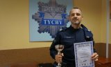 Obrazek przedstawia umundurowanego policjanta, który trzyma pucha oraz dyplom, w tle na ścianie widoczne logo Komendy Miejskiej Policji w Tychach