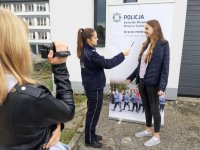 Obrazek przedstawia zajęcia praktyczne nagrywania się przed kamerą, widoczna umundurowana policjantka oraz młoda dziewczyna na tle banera z Komendy Miejskiej Policji w Tychach. Po lewej stronie widoczna druga dziewczyna, która kameruje