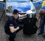 Obrazek przedstawia umundurowane policjanta który stoi przed oznakowanym radiowozem z dzieckiem, który ma założony strój PZ, który wykorzystywany jest do zabezpieczenia meczy