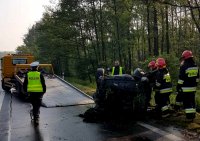 Obrazek przedstawia strażaków przy samochodzie, który &quot;dachował&quot;  oraz policjanta i lawetę