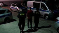 Na obrazku widać policjantów prowadzących zatrzymanego, widoczne zaparkowane policyjne radiowozy