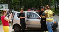 Obrazek przedstawia dwóch strażaków, za nimi samochód, w tle widoczni policjanci, obok nich dwie kobiety, jedna z n9ich zakłada mundur strażaka