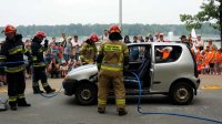 Na zdjęciu strażacy wycinają szybę w samochodzie, widoczne wokół zgromadzone licznie dzieci wraz ze swoimi opiekunami