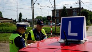Policjant z funkcjonariuszem straży ochrony kolei rozmawiają z kursantem nauki jazdy przed przejazdem kolejowym