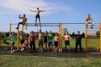 Zdjęcie przedstawia grupę osób na siłowni na świeżym powietrzu,część z nich jest bez koszulki po prawej stroni stoi z nimi dzielnicowy w mundurze, trzech z mężczyzn znajduje się na drabinkach.