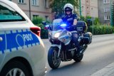 Obrazek przedstawia kawałek radiowozu, a za nim policjanta na motocyklu