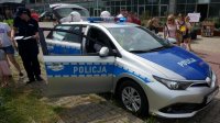 Na zdjęciu widoczny radiowóz oraz policjantka, która rozdaje dzieciom kolorowanki, w tle widoczne inne osoby