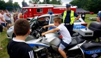 Na zdjęciu widoczny policjant, za nim stoi radiowóz, przed nim z kolei stoi policyjny motocykl, na którym siedzi chłopiec, wokół widoczni inni uczestnicy festynu