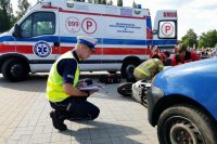 Na zdjęciu policjant, który wykonuje czynności podczas symulacji wypadku i dokumentuje zdarzenie, klęczy przed motocyklem, w tle widoczna karetka pogotowia