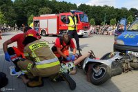 Na zdjęciu widoczni ratownicy, którzy udzielają pomocy poszkodowanemu podczas symulacji wypadku, obok policjant, widoczni przyglądający się temu uczestnicy festynu, w tle widoczny wóz straży pożarnej