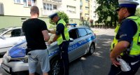 Przy radiowozie widoczny policjant z mężczyzną, dale drugi policjant asekuruje czynności legitymowania.