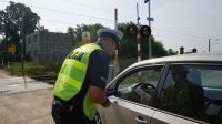 umundurowany policjant w kamizelce odblaskowej, na plecach napis POLICJA rozmawia z kierowcą jasnej osobówki w tle widać tory i biało czerwony szlaban