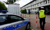 Na zdjęciu widoczny policjant z drogówki w żółtej kamizelce, stojący przed Komenda Miejska Policji w Tychach, obok stoi zaparkowany radiowóz oraz biały samochód.