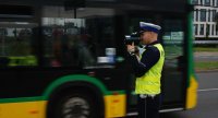 Na zdjęciu widoczny policjant wydziału ruchu drogowego w kamizelce odblaskowej stoi bokiem, w tle przejeżdża autobus.