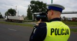 Na zdjęciu widoczny policjant wydziału ruchu drogowego w kamizelce odblaskowej stoi tyłem na drodze, w tle widoczny samochód ciężarowy.