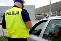 Na zdjęciu widoczny policjant wydziału ruchu drogowego w kamizelce odblaskowej stoi przy zatrzymanym pojeździe i rozmawia z kierującym.