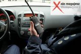 Na zdjęciu widoczne umundurowany policjant siedzący wewnątrz radiowozu i trzymający radiostację.