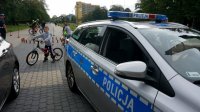 Na zdjęciu widoczny radiowóz, dalej dwóch rowerzystów, po lewej stronie policjant, który przygląda się jeździe rowerzysty.