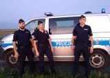 Trzech umundurowanych policjantów ( w  środku policjantka) stoi przed oznakowanym radiowozie.