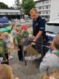 Umundurowany dzielnicowy trzyma tarcze z napisem &quot;POLICJA&quot; wokół niego stoją dzieci.