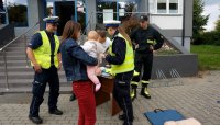 Kobieta z dzieckiem na rękach stoi przy stoliku z gadżetami, policjantka daje kobiecie odblaski. Obok widoczny strażak oraz policjant oraz mężczyzna ze Stowarzyszenia Sportowego NOL Tychy.