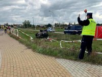 Na zdjęciu widoczni policjanci wydziału ruchu drogowego w żółtych kamizelkach, którzy wykonują pomiary w związku ze śmiertelnym wypadkiem. Dalej po prawej strony widoczne dwa radiowozy oraz wydachowany samochód w rowie.