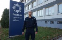 Umundurowany policjant stoi przed budynkiem Komendy Miejskiej Policji w Tychach przy banerze komendy.