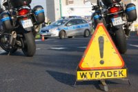 na pierwszym planie jest znak żółty trójkąt z czarnym wykrzyknikiem i napisem WYPADEK
w tle widać dwa motocykle policyjne oraz oznakowany radiowóz policyjny
wszystko stoi na czarnej asfaltowej drodze