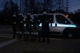 Na zdjęciu widoczni umundurowani policjanci, którzy zabezpieczają mecz piłki nożnej. Pięciu funkcjonariuszy stoi przed radiowozem w kaskach, a trzech z nich trzyma tarcze z napisem &quot;POLICJA&quot;.