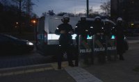 Na zdjęciu widoczni umundurowani policjanci, którzy zabezpieczają mecz piłki nożnej. Pięciu funkcjonariuszy stoi przed radiowozem w kaskach, a trzech z nich trzyma tarcze z napisem &amp;quot;POLICJA&amp;quot;.