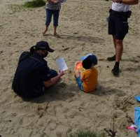 Policjanta siedzi na plaży z chłopcem i przekazuje mu kolorowankę.