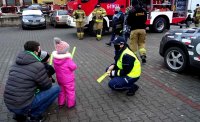 Policjantka rozmawia z mężczyzną oraz dziećmi, rozdaje odblaski, w tle strażacy i wóz strażacki.