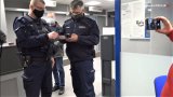 Na zdjęciu widoczni dwaj umundurowani policjanci, jeden trzyma dokument, drugi telefon. W tle widoczny mężczyzna. Z prawej strony widoczna ręka z telefonem który ma włączoną kamerę.